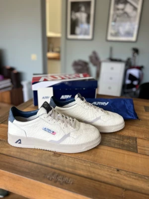 Vita Autry sneakers  - Säljer ett par vita sneakers från Autry med blå detaljer på häl och plös. Skorna har en låg silhuett, klassisk snörning och är tillverkade i strukturerat läder. Snygg logga på sidan och bekväm innersula. Perfekta för en clean och stilren look.