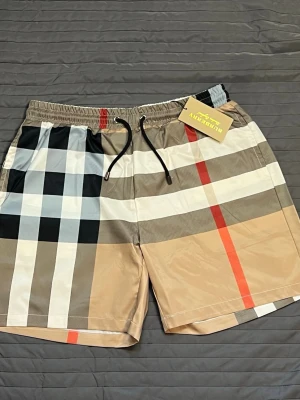 Burberry shorts - Snygga badshorts från Burberry med klassiskt rutigt mönster i beige, svart, vit och röd. Shortsen har elastisk midja med svart snörning och en bakficka. Perfekta för sommaren och strandhäng.