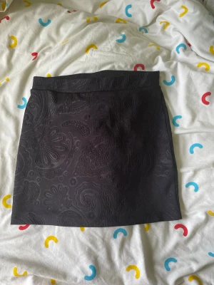 Svart kortkjol från Vero Moda - Snygg svart kortkjol från Vero Moda med diskret paisleymönster i tyget. Kjolen har en rak passform och är tillverkad i ett glansigt material som ger en elegant känsla. Perfekt att matcha med en enkel topp eller stickad tröja.