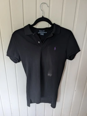 Svart pikétröja från Ralph Lauren - Klassisk svart pikétröja från Ralph Lauren med korta ärmar och krage. Tröjan har två knappar framtill och den ikoniska lilla loggan broderad i lila på bröstet. Perfekt till jeans eller kjol för en clean och stilren look.