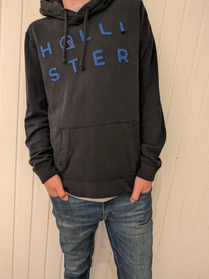 Svart hoodie från Hollister - Snygg svart hoodie från Hollister med blå broderad logga över bröstet. Klassisk känguruficka framtill och justerbar huva med snören. Perfekt för en avslappnad och trendig stil. Tillverkad i mjukt material som är skönt att bära.