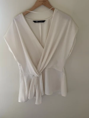 Vit omlottopp från Zara - Stilren vit omlottopp från Zara med v-ringning och knyt i midjan. Toppen har korta ärmar och ett mjukt, lätt material som ger ett snyggt fall. Perfekt för dig som gillar minimalistisk och fräsch stil.