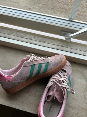 Adidas rosa och gröna sneakers - Säljer ett par snygga Adidas sneakers i rosa mocka med gröna ränder och detaljer. Skorna har rosa insida och häl, samt klassisk gummisula. Perfekta för dig som vill sticka ut med färg och retrostil. Låga och med snörning för skön passform.
