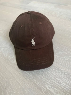 Brun keps från Polo Ralph Lauren - Snygg brun keps från Polo Ralph Lauren med klassisk böjd skärm och broderad logga framtill. Justerbart spänne bak och 'POLO' broderat i vitt. Tillverkad i bomullstyg som känns både slitstarkt och bekvämt.