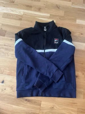 Mörkblå zip hoodie från Fila - Snygg mörkblå hoodie från Fila med dragkedja framtill och vit rand över bröstet. Fila-logga på vänster sida. Tröjan har hög krage, långa ärmar och två fickor. Perfekt för en sportig och avslappnad stil.