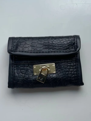 Svart plånbok med krokopräglat mönster - Snygg svart plånbok i konstläder med krokopräglat mönster och guldfärgat lås framtill. Plånboken har en vikbar design och dragkedjefack på insidan. Perfekt för dig som gillar stilrena detaljer och trendiga accessoarer.😊