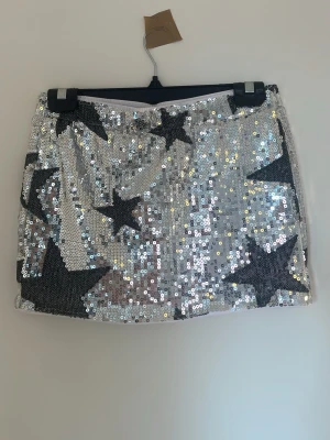 Glittrig silver skort med stjärnor - Kort kjol täckt av silverfärgade paljetter och stora svarta stjärnor med inbyggda shorts. Kjolen är i storlek S från SHEIN. Använd en gång.
