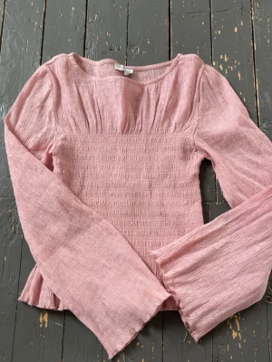 Ljusrosa blus med långa ärmar - Supersöt ljusrosa blus med smockad midja och vida långa ärmar. Tillverkad i ett tunt, krinklat bomullsliknande material som ger en luftig känsla. Perfekt för vår och sommar när du vill ha något lätt och fräscht. Storlek xs och använd ett par gånger då det är en fläck i ärmen och ett litet hål som knappt syns❣️