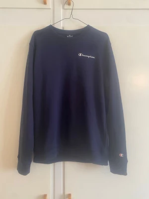 Mörkblå sweatshirt från Champion - Mörkblå sweatshirt från Champion med klassisk logga broderad på bröstet och ärmen. Tröjan har rund halsringning, långa ärmar och ribbade muddar vid ärmslut och nederkant. Perfekt för en avslappnad och sportig stil.
