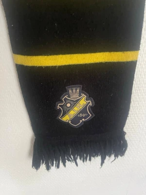 AIK halsduk svart och gul - Säljer en klassisk AIK halsduk i svart och gult med klubbens logga broderad på ena änden. Halsduken har breda ränder och fransar i ändarna, perfekt för att visa ditt stöd på matchen eller i vardagen.