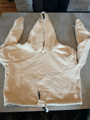 Beige hoodie med dragsko - Säljer en beige hoodie med huva och dragsko i nederkanten. Hoodien har långa ärmar och en relaxed passform. Materialet ser ut att vara mjukt bomullstyg, perfekt för chill dagar. Liten patchdetalj nere till vänster.