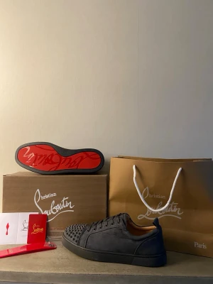 Gråa Louboutin skor  - Säljer ett par svarta sneakers med coola nitar på tån och den ikoniska röda sulan. Skorna har en låg profil och är tillverkade i mockaliknande material med svarta skosnören. Perfekt för dig som vill sticka ut med din stil.