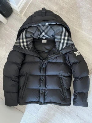 Märkes jacka/väst med huva - Snygg svart dunjacka med stor huva och rutigt grått/vitt foder. Jackan har två dragkedjeförsedda fickor framtill, knappar och dragkedja för stängning samt en patch på vänster ärm. Puffig modell som håller dig varm under kalla dagar. Skick 10/10