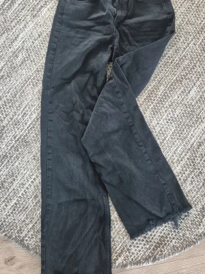 Svarta raka jeans från Pull&Bear - Säljer ett par svarta jeans från Pull&Bear i rak modell med rå kant nertill. Jeansen har hög midja, klassisk femficksdesign och dragkedja med knapp. Perfekta för en avslappnad och trendig look.