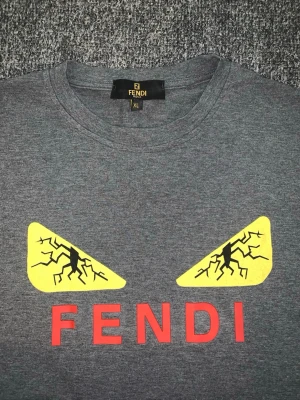 Grå Fendi t-shirt med tryck - XL men sitter som L Snygg grå t-shirt från Fendi med ikoniskt ögon-tryck i gult och svart samt röd FENDI-text på bröstet. Klassisk rund hals och korta ärmar. Perfekt för dig som vill sticka ut med en cool och trendig look.