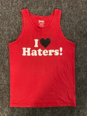 Röd DGK linnetopp med tryck - Säljer en röd linnetopp från DGK med det ikoniska trycket 'I ♥ Haters!' på bröstet. Toppen är ärmlös och gjord i 100% bomull, perfekt för en chill och streetinspirerad stil.