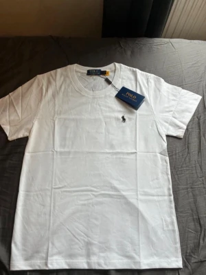 Vit t-shirt från Polo Ralph Lauren - Klassisk vit t-shirt från Polo Ralph Lauren med rund hals och korta ärmar. T-shirten har den ikoniska broderade loggan på bröstet och är gjord i mjuk bomull för en skön känsla. Perfekt basplagg till garderoben. Storlek S