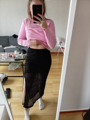 Svart långkjol med slits - Säljer en svart långkjol i virkad stil med hög slits på ena sidan. Kjolen har ett snyggt genombrutet mönster och sitter tajt över höfterna. Perfekt för dig som vill ha en trendig och edgy look.