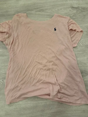 Ljusrosa t-shirt från Polo Ralph Lauren - Säljer en ljusrosa t-shirt från Polo Ralph Lauren med klassisk blå logga broderad på bröstet. T-shirten är kortärmad och gjord i mjuk bomull, perfekt för en avslappnad och stilren look.
