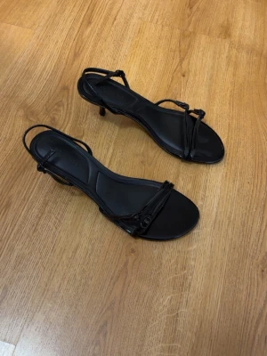 Svarta remsandaler med smal klack - Snygga svarta sandaler med tunna remmar och smal klack. Öppen tå och justerbar rem runt hälen. Tillverkade i ett glansigt material som ger en elegant look. Perfekta för dig som gillar stilrena och trendiga skor. Vid intresse tveka inte hör av dig. Mvh 