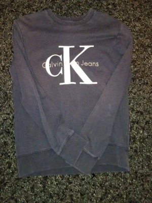 Mörk blå sweatshirt från Calvin Klein - Snygg mörkblå sweatshirt från Calvin Klein Jeans med stort vitt CK-tryck på bröstet. Tröjan har rund halsringning och långa ärmar. Perfekt för en avslappnad och trendig look.