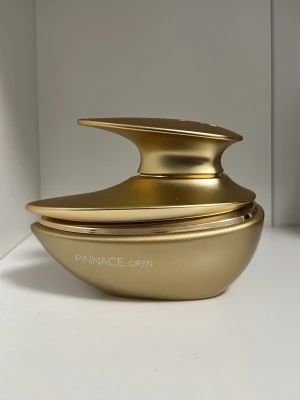 Pinnace Oryn av french avenue  - Unik parfymflaska i metalliskt guld med futuristisk, asymmetrisk form. Flaskan har elegant design och texten 'PINNACE ORYN' på sidan. Perfekt som lyxig detalj på sminkbordet eller i badrummet.