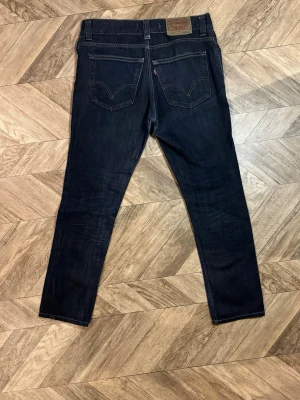 Levi's 511 mörkblå jeans - Säljer ett par mörkblå Levi's 511 jeans med klassisk femficksdesign och raka ben. Jeansen har snygga kontrastsömmar och den ikoniska läderlappen bak i midjan. Perfekta för dig som gillar en tidlös och clean look. Längd 99cm bredd 42cm.