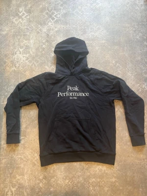 Svart hoodie från Peak Performance - Snygg svart hoodie från Peak Performance med vit logga och text 'Est. 1986' på bröstet. Klassisk känguruficka framtill och mjuk insida i fleece. Huva med dragsko och ribbade muddar vid ärmar och nederkant. Perfekt för en avslappnad och sportig stil. Den är nött i armbågen och vid handlederna