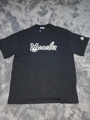 Svart Moncler t-shirt med logga - Snygg svart t-shirt från Moncler med stor vit broderad logga på bröstet och klassisk rund hals. T-shirten har korta ärmar och en liten Moncler-patch på ena ärmen. Perfekt för dig som gillar streetwear och vill ha något stilrent. Passar oversized.