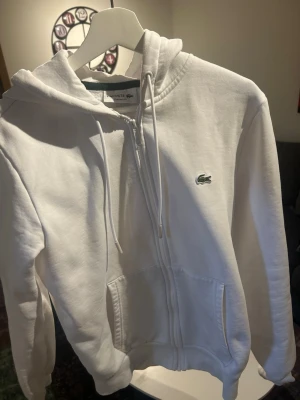 Vit zip hoodie från Lacoste - Säljer en vit hoodie från Lacoste med dragkedja framtill och klassisk huva med dragsko. Hoodien har två fickor och den ikoniska gröna krokodilloggan på bröstet. Modellen är Classic Fit och materialet känns mjukt och bekvämt.