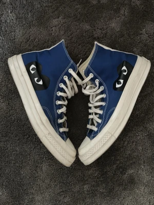 Blå Converse x CDG Play Highs - Säljer ett par blå Converse x Comme des Garçons Play high top sneakers med vita sulor och snören. Skorna har det ikoniska CDG Play-hjärtat på sidan och klassisk Converse-stjärna på insidan. Tillverkade i canvas med gummisula och metallöljetter.