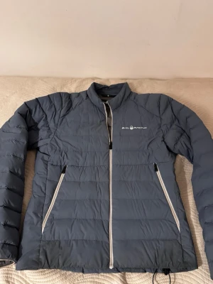 Blå dunjacka från Sail Racing - Snygg blå dunjacka från Sail Racing med vita dragkedjor och logga på bröstet. Jackan har två snedställda fickor framtill och quiltad design. Perfekt för kalla dagar när du vill hålla värmen och ändå se stilren ut.