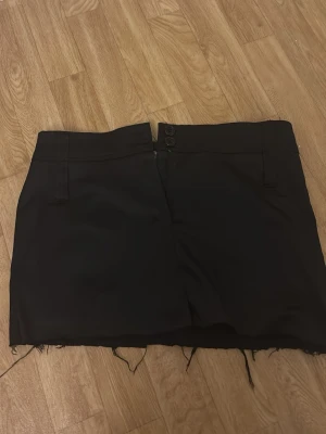 Svart kort kjol med rå kant - Stilren svart kjol med låg midja, två knappar framtill och rå, fransig kant nedtill. Kjolen har fickor både fram och bak med dekorativa lock och knappar bak. Perfekt för en edgy och trendig look. Och är i kostym tyg 