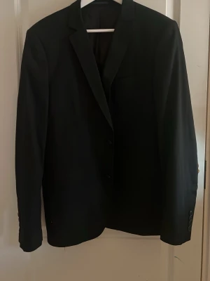 Svart kavaj från Zara Man - Stilren svart kavaj från Zara Man Basic med klassisk passform och tvåknäppning. Kavajen har smala slag, innerfoder och knappdetaljer vid ärmslut. Perfekt för en clean och modern look.