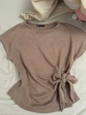 topp med knyt Zara - Säljer en beige topp med korta ärmar och snygg knytning i midjan. Toppen har en enkel och stilren design som passar perfekt till jeans eller kjol. Materialet känns mjukt och bekvämt mot huden.