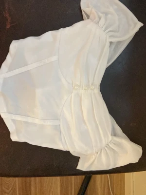 Vit axelbandslös topp med puffärm - Supersöt vit axelbandslös topp med stora puffärmar och pärldetaljer framtill. Toppen har en bandeauformad överdel och är gjord i ett tunt, lätt material som känns luftigt. Perfekt för dig som vill sticka ut med en trendig och feminin look.