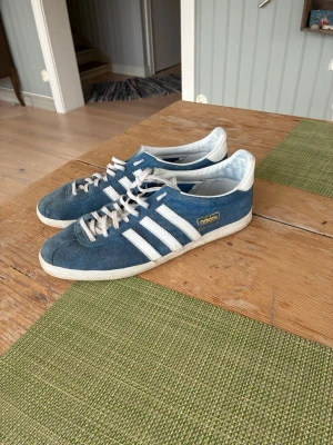 Adidas Gazelle blå sneakers - Säljer ett par klassiska Adidas Gazelle sneakers i blå mocka med vita detaljer och de ikoniska tre ränderna på sidan. Skorna har vit sula och vita skosnören samt guldfärgad Adidas-logga på sidan. Perfekta för en stilren och sportig look. Passar 42-43