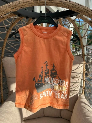 Linnetopp strl. 104 - Orange ärmlös linnetopp från tutta med coolt piratskepp-tryck och texten 'Monsters of Seven Seas' framtill. Tillverkad i 100% bomull, perfekt för varma dagar och en avslappnad stil.
