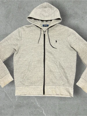 Grå zip hoodie från Polo Ralph Lauren - Snygg gråmelerad hoodie med dragkedja från Polo Ralph Lauren. Klassisk huva med svarta snören, två fickor fram och ikoniska loggan broderad på bröstet. Tillverkad i mjukt bomullsmaterial som känns skönt mot huden. Perfekt till chill eller streetwear.