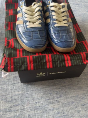 Adidas Wales Bonner croc - Unika blå Adidas sneakers med krokodilpräglat mönster och klassiska vita ränder på sidorna. Skorna har vita snören, gulddetaljer vid snörningen och beige sula. Tungan har ett mönster i vitt och gult som ger extra karaktär. Perfekta för dig som vill sticka ut.