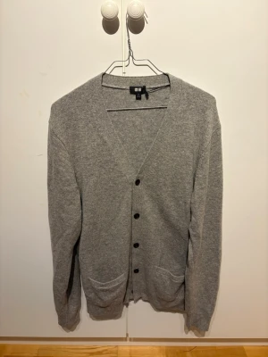Grå kofta från Uniqlo, M - Klassisk grå kofta från Uniqlo med v-ringning och knäppning framtill. Tillverkad i mjukt material och har två fickor framtill. Perfekt för lager på lager och enkel att matcha med olika outfits.