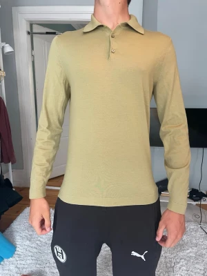 Olivgrön långärmad pikétröja - Snygg olivgrön långärmad pikétröja med klassisk krage och tre knappar framtill. Tröjan har en smal passform och är tillverkad i ett tunt, mjukt material som känns skönt mot huden. Perfekt för dig som gillar stilrena och enkla plagg.