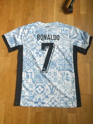 Ronaldo fotbollströja - Säljer en snygg fotbollströja med Ronaldo och nummer 7 på ryggen. Tröjan har ett unikt ljusblått och vitt mönster med inslag av olika symboler och svarta sidopaneler. Perfekt för dig som vill sticka ut på planen eller visa ditt stöd för Ronaldo.