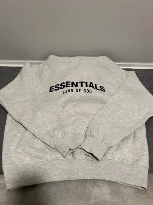 Grå Essentials Fear of God hoodie - Snygg ljusgrå hoodie från Fear of God Essentials med svart tryck på bröstet. Klassisk passform med ribbade muddar och mjukt material som känns riktigt skönt. Perfekt för dig som gillar streetwear och vill ha en clean look.