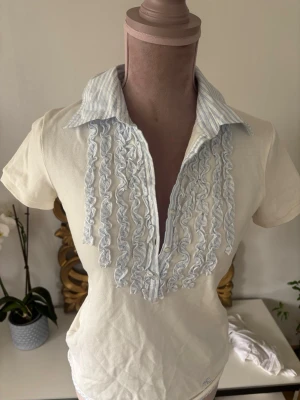 Vit blus med volangdetaljer MaxMara  - Snygg vit blus med korta ärmar och ljusblå volangdetaljer framtill. Kragen har ett klassiskt skjortutseende med tunna blå ränder. Blusen har en feminin och elegant look, perfekt till jeans eller kjol. Materialet känns mjukt och lätt.