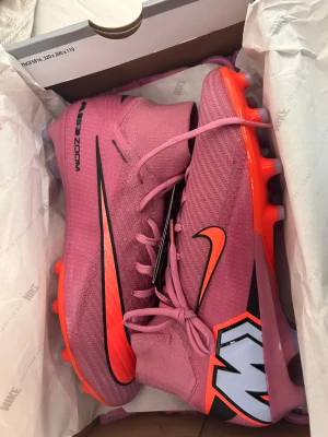 Superfly 10 gripknit - Säljer ett par grymma rosa Nike fotbollsskor med högt skaft och orangea detaljer. Skorna har svart Nike-logga, snörning och en robust sula med dobbar för bra grepp på planen. Perfekta för dig som vill sticka ut på matchen! Helt ny med lappar
