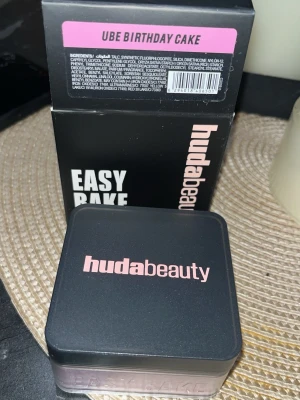 Huda Beauty Easy Bake Ube Birthday Cake - Lös foundation i puderform från Huda Beauty i färgen Ube Birthday Cake. Kommer i en fyrkantig burk med svart lock och rosa text. Perfekt för att fixera sminket och ge en jämn finish. Förpackningen är stilren och modern.