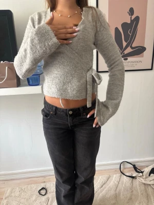 Grå omlottstickad tröja - Mysig grå stickad omlottopp med långa ärmar och knytband i sidan. Tröjan har en mjuk, fluffig känsla. Perfekt att styla med höga jeans för en trendig look.
