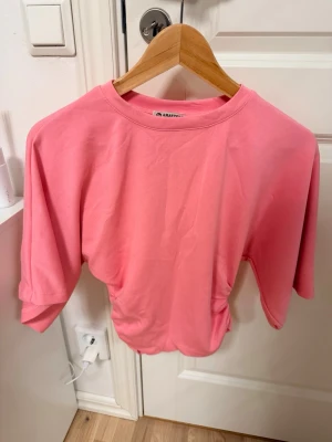 Rosa t-shirt från Amayteks - Snygg rosa t-shirt från Amayteks i storlek M. T-shirten har rund halsringning, korta ärmar och en lätt croppad passform med rynkade detaljer i sidorna. Perfekt för en trendig och avslappnad stil. Passar S/M det är exakt samma tröja som den gula. Så du kan se hur den sitter på i sista bilden!