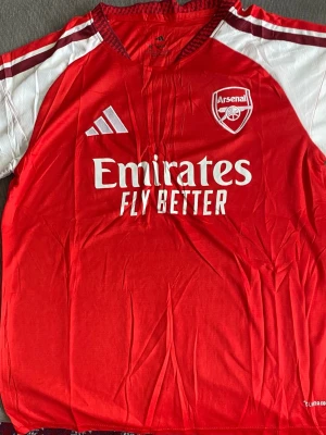 Arsenal matchtröja röd Adidas M - Säljer en röd Arsenal matchtröja från Adidas i storlek M. Tröjan har vita detaljer på ärmarna, klubbmärke och Adidas-logga på bröstet samt sponsortryck 'Emirates Fly Better' framtill. Tillverkad i lätt och ventilerande material, perfekt för fotboll eller träning. M passar till S